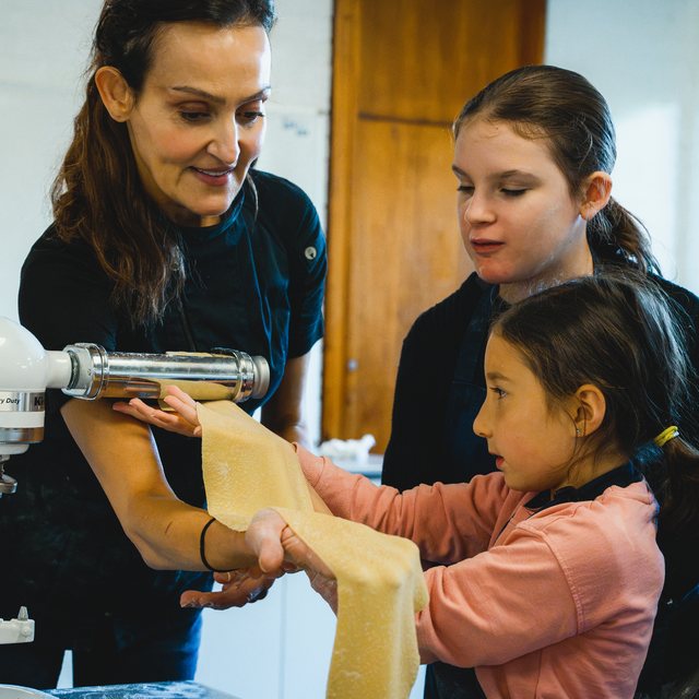 Yvonne guiding two girls as they feed a sheet of fresh pasta through the roller