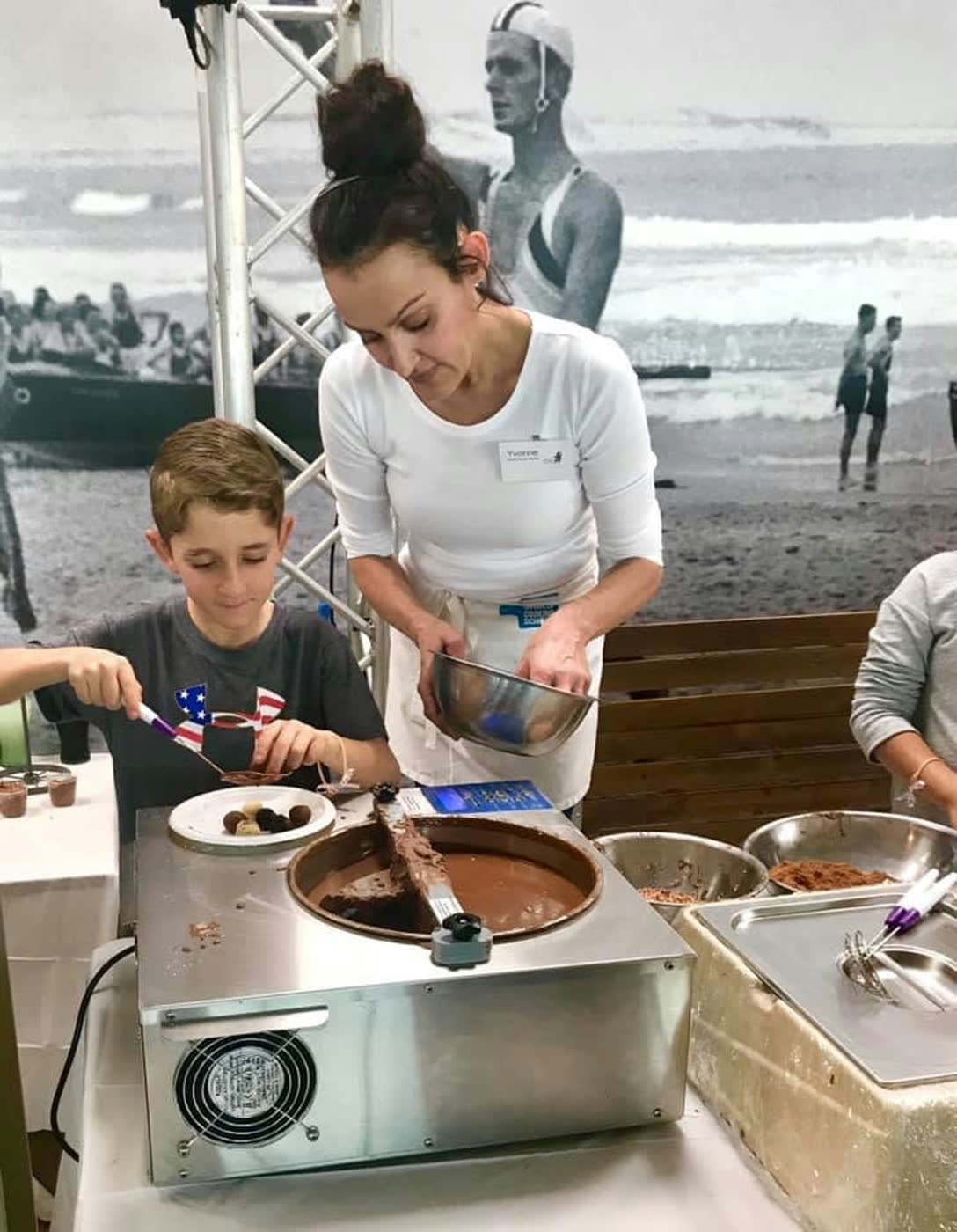 A boy working chocolate on the tempering machine while Yvonne steadies the tray