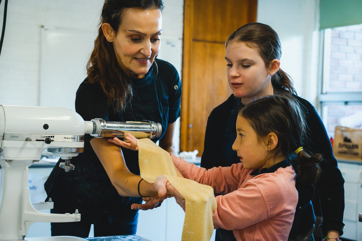 Yvonne feeding a pasta sheet through the machine with two young helpers