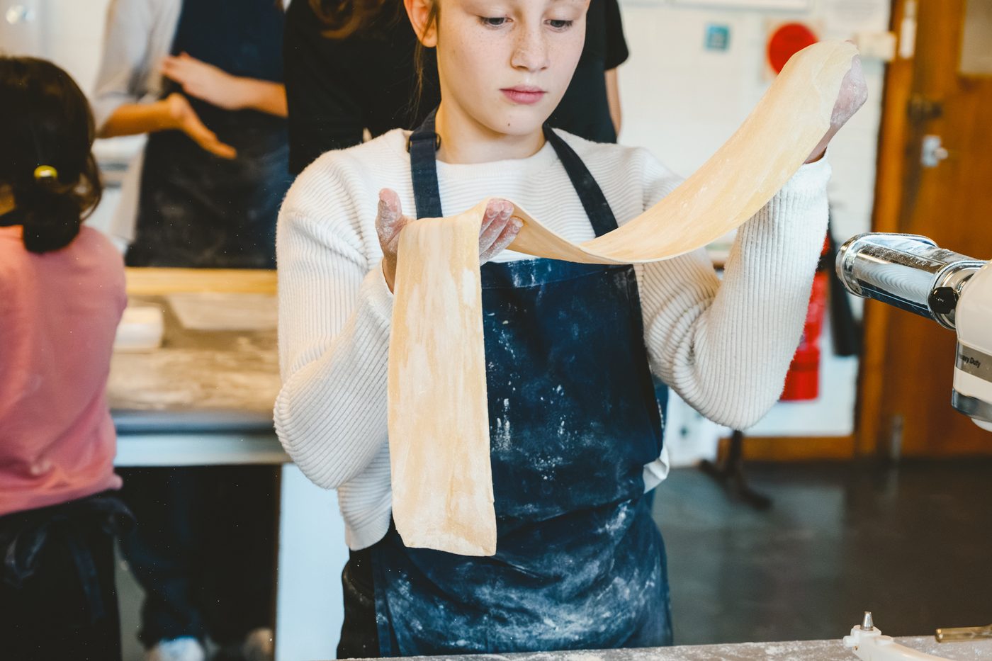 A student in a floury apron stretching a wide sheet of fresh pasta