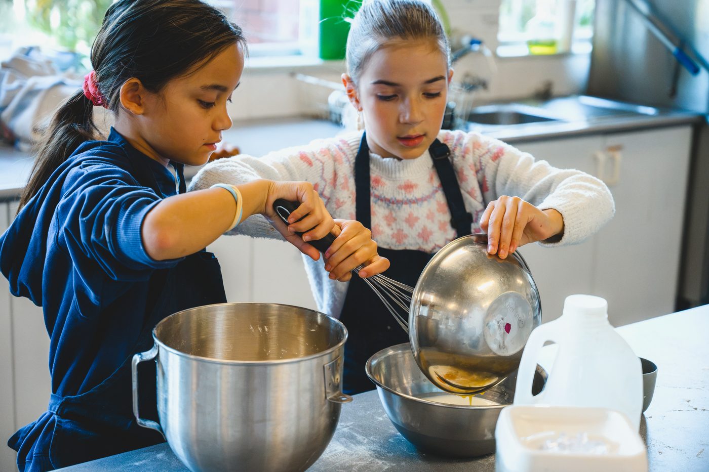 Two girls whisking together, one steadying the bowl while the other pours
