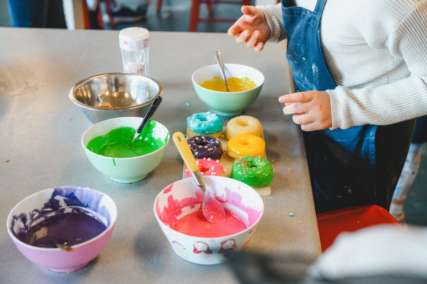 Bowls of brightly coloured icing beside a board of freshly decorated doughnuts