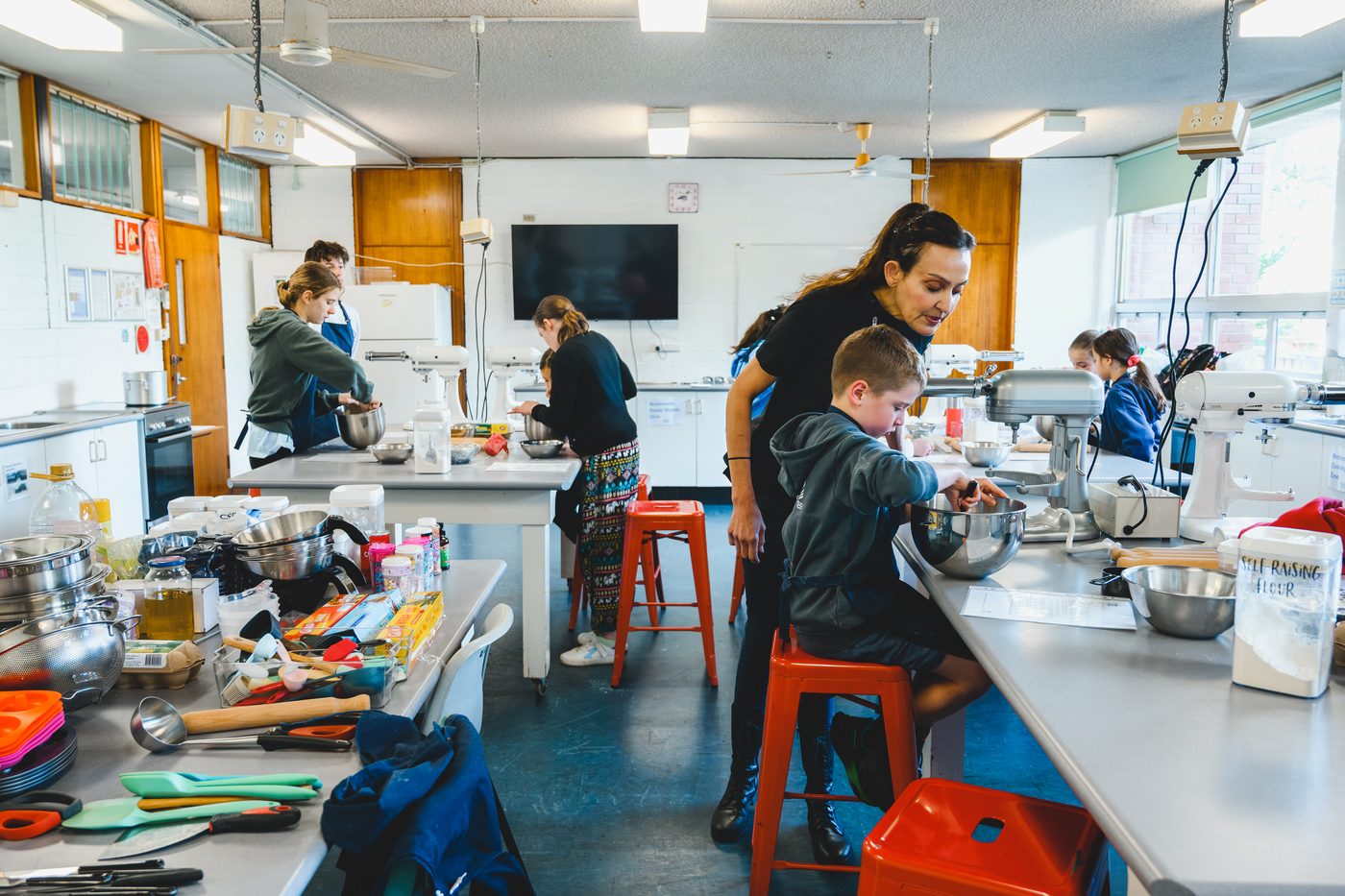 A busy class kitchen with students at every bench, and Yvonne helping a young boy at the mixer