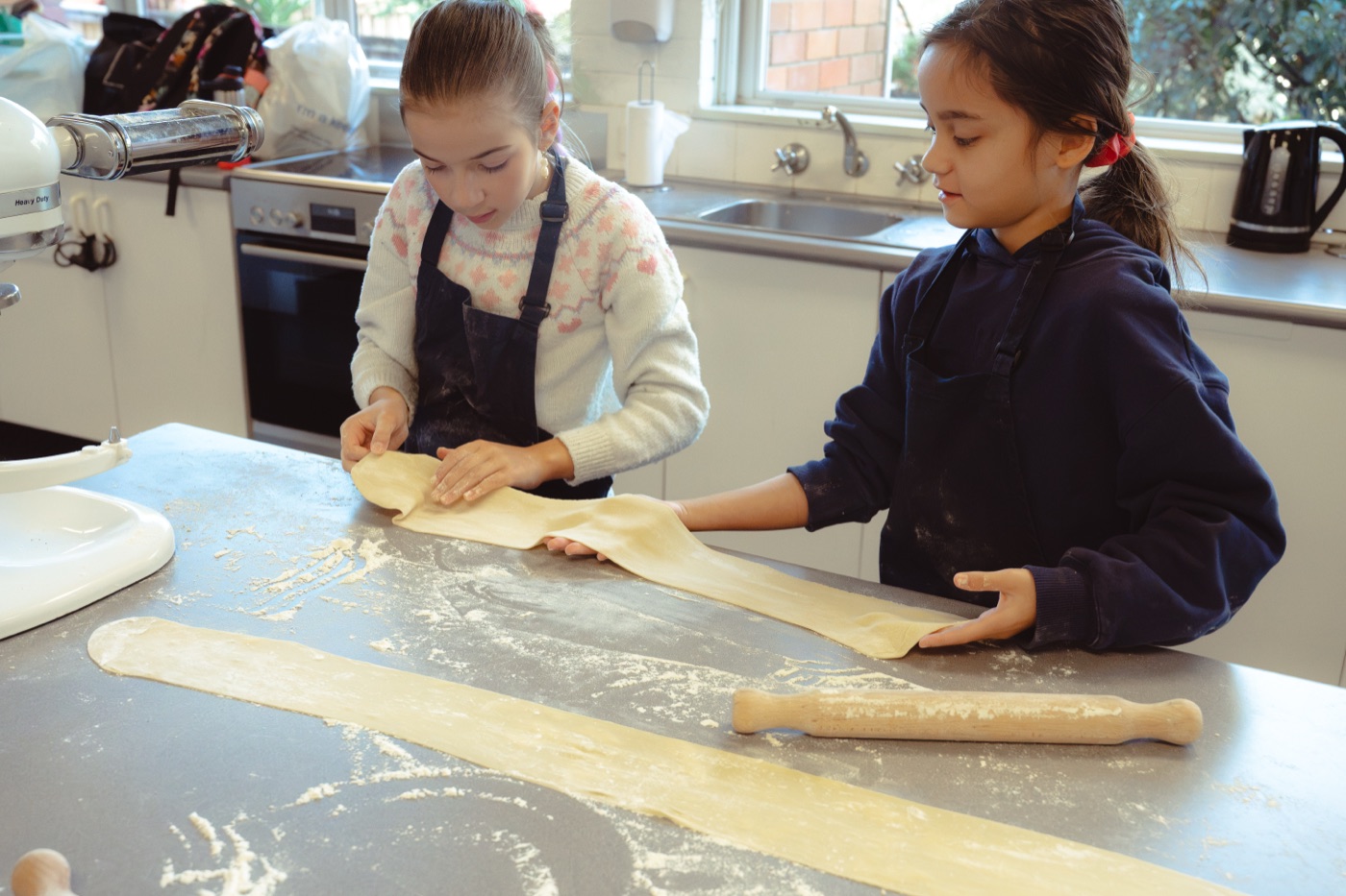 Two girls stretching a long sheet of fresh pasta across a floured bench