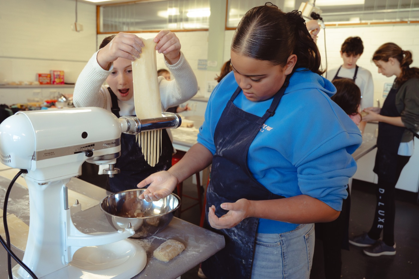 One student lifting a pasta sheet from the roller while another works beside her