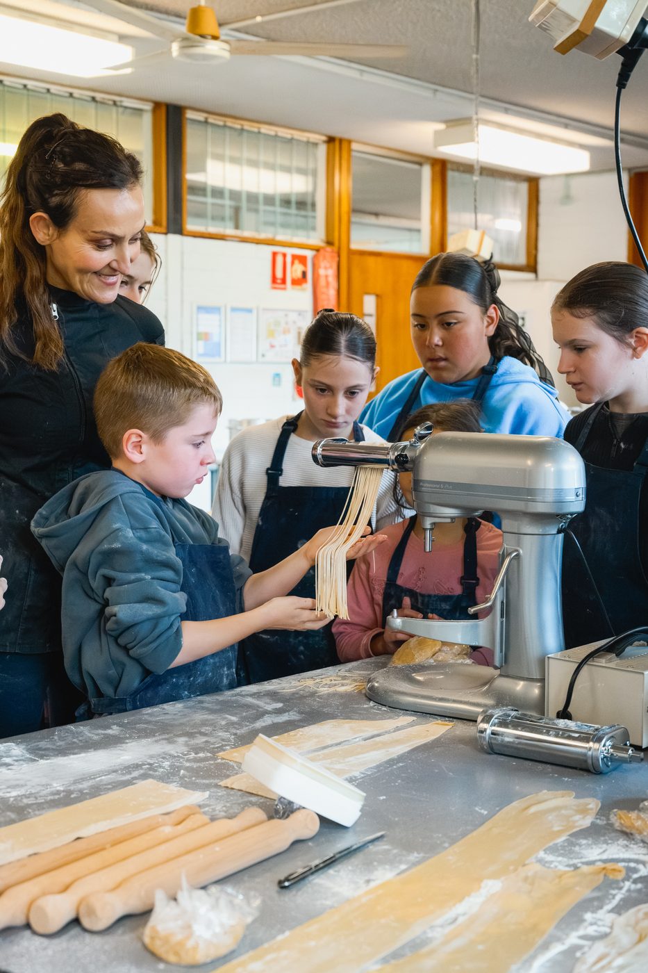 Yvonne guiding a young student at the pasta machine while the class looks on