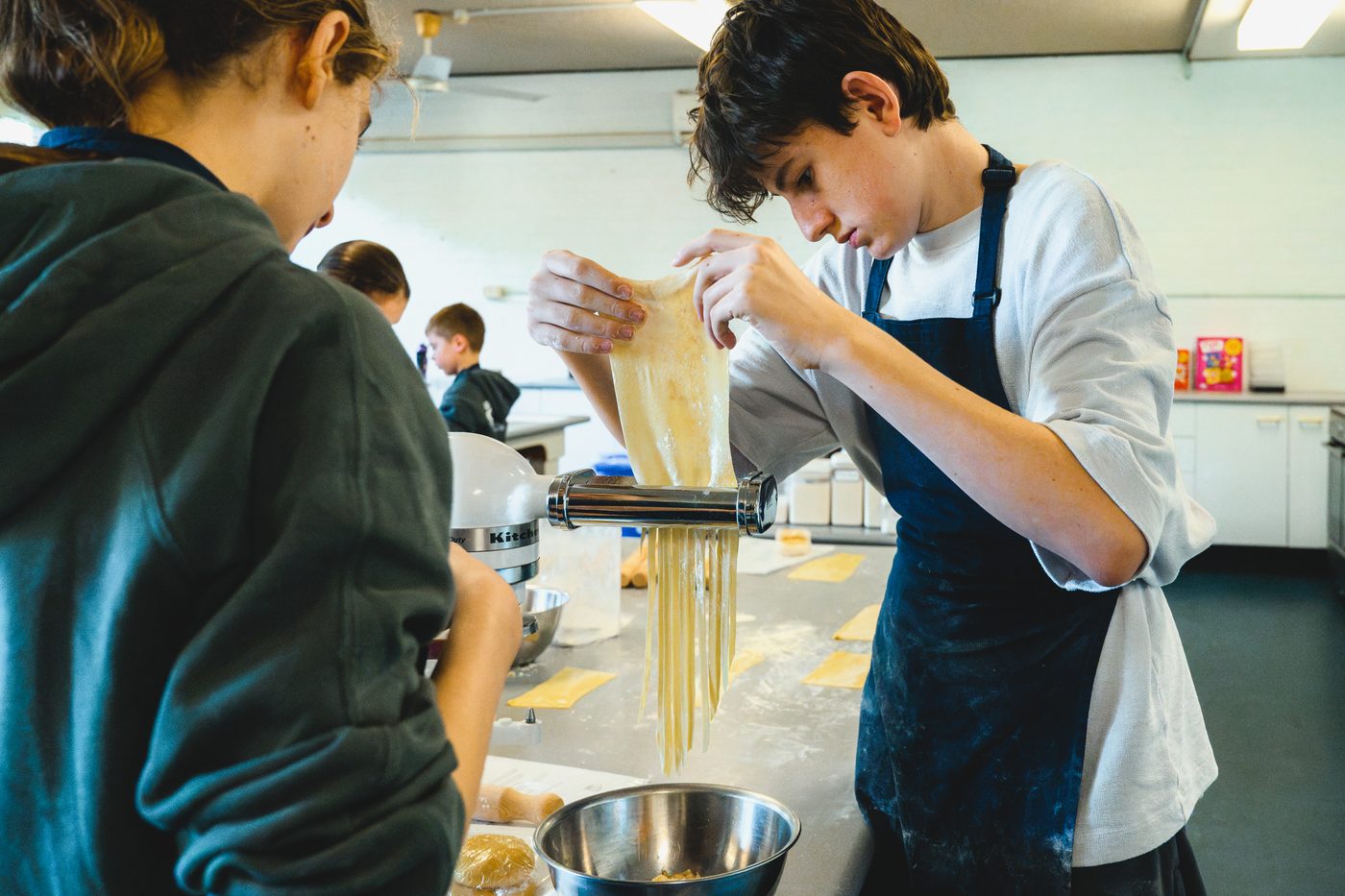 Two boys guiding fresh spaghetti out of the pasta machine into a bowl