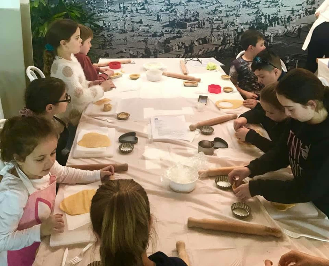 A long cloth-covered table of children rolling dough and pressing out shapes with cutters