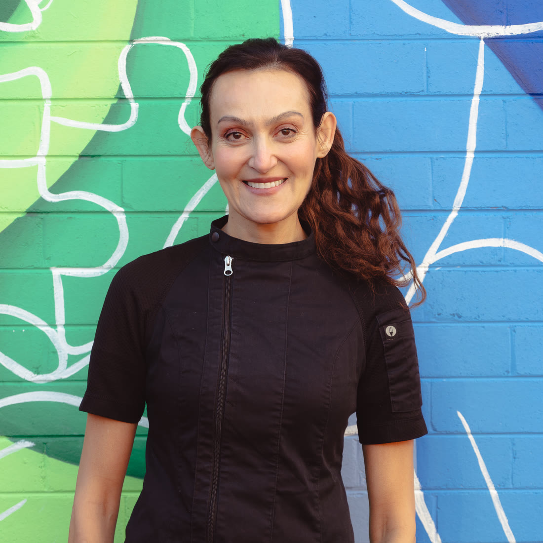 Yvonne, qualified chef and founder of Sutherland Shire Cooking School, in her chef's jacket in front of a brightly painted mural